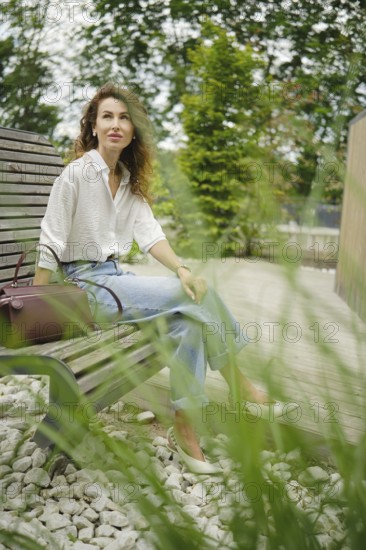 A woman sits calmly on a wooden garden bench, dressed in a light shirt and jeans. Tall grass sways around her as she gazes thoughtfully into the distance on a tranquil afternoon