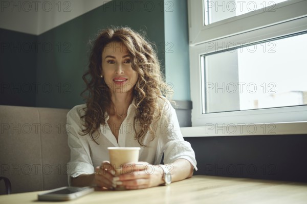 A woman with curly hair sits at a wooden table in a cafe, holding a cup of coffee. She smiles while looking out of a large window that lets in bright afternoon light