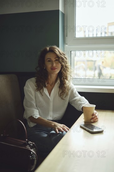 A woman with curly hair sits at a wooden table in a cafe, holding a paper cup. Sunlight streams through the window, illuminating her casual outfit as she seems relaxed and content