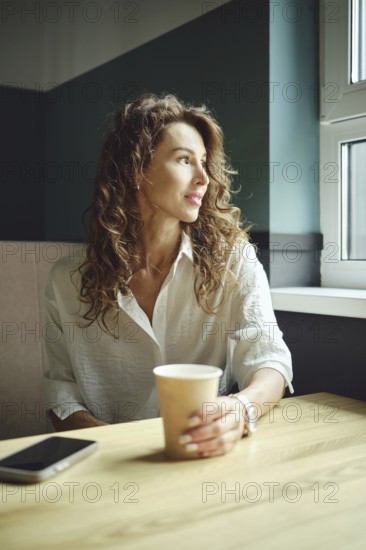 A woman with curly hair sits at a wooden table in a cozy cafe, holding a coffee cup. She gazes out the window, lost in thought, as natural light brightens her surroundings