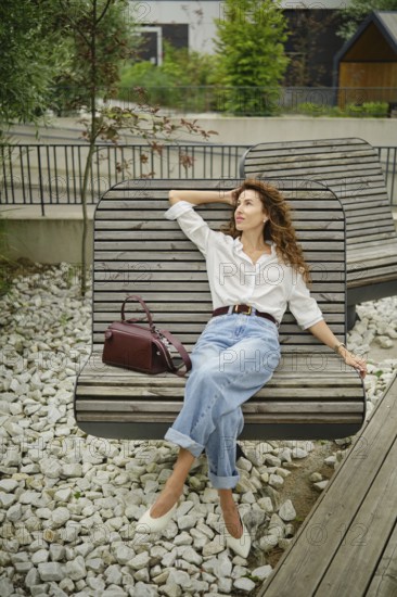 A woman enjoys her time on a stylish bench in a contemporary outdoor area filled with greenery. She wears a white shirt and high-waisted jeans, looking relaxed while holding a handbag
