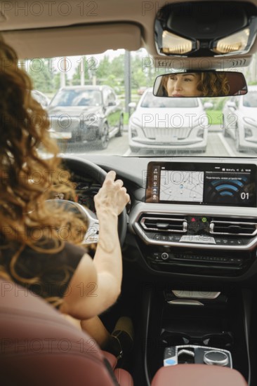 A woman with curly hair is focused on driving a car, checking the navigation screen. She is inside a modern vehicle parked among several other cars on a clear day, AI generated
