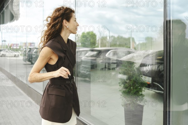 A woman stands beside a large glass window in a car showroom, gazing at displayed vehicles. Her stylish outfit adds to the contemporary atmosphere of the space during daytime, AI generated