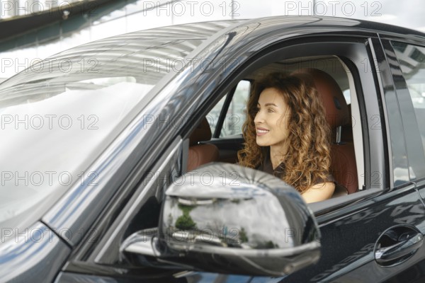 A woman with curly hair is smiling as she sits in the driver's seat of a sleek car. The vehicle is parked next to a contemporary building with large windows, reflecting the surroundings, AI generated