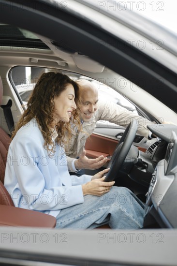 A woman sits in the driver's seat of a car, looking at the dashboard screen, while an older man leans in from the opened door, explaining features and options, AI generated