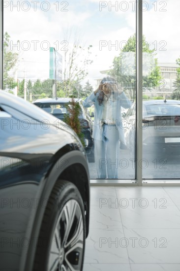A woman stands outside looking in through large glass windows to a car dealership, admiring various cars displayed. The atmosphere is bright and inviting with a clear sky above, AI generated