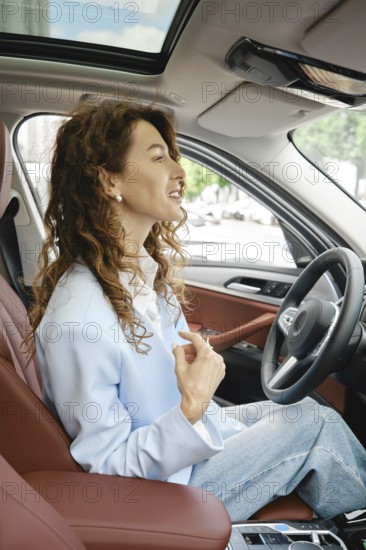 A woman with curly hair is seated in the driver's seat of a modern car, smiling and looking forward. Sunlight pours through the windows, enhancing her cheerful expression, AI generated