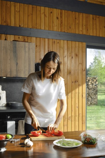 A woman is chopping fresh bell pepper in a rustic kitchen. Sunlight pours through the window, highlighting the wooden walls and the outdoor greenery