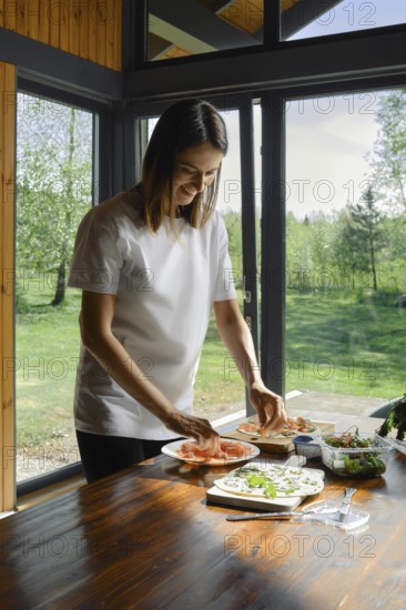 A woman arranges slices of salmon over tortilla on a wooden table in a bright kitchen. Sunlight streams through large windows, highlighting the lush greenery outside, creating a warm atmosphere