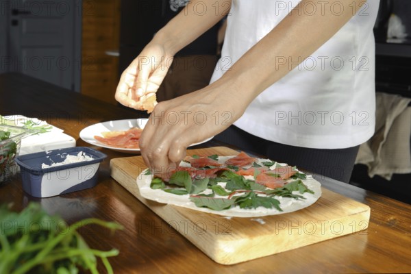 A person is preparing a healthy dish in a cozy kitchen setting. Fresh greens, slices of salmon, and creamy toppings are arranged on a flatbread, emphasizing wholesome cooking at home