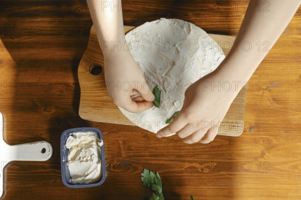 Hands carefully place fresh herbs onto a creamy spread during appetizer preparation. A container of cream cheese sits nearby on a warm wooden surface, showcasing a moment of healthy cooking