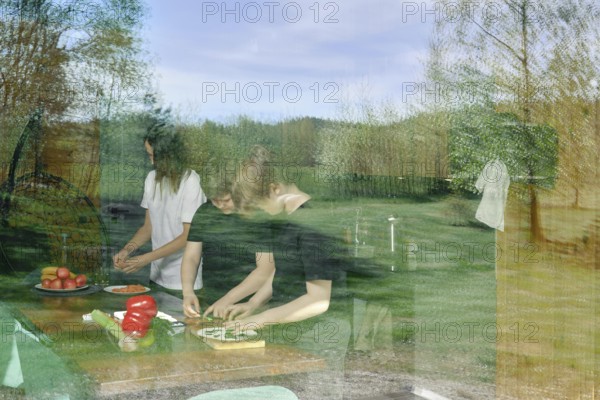 The view through the window to the kitchen, where the family is preparing a light snack. The glass reflects the meadow and the forest where the country house is located