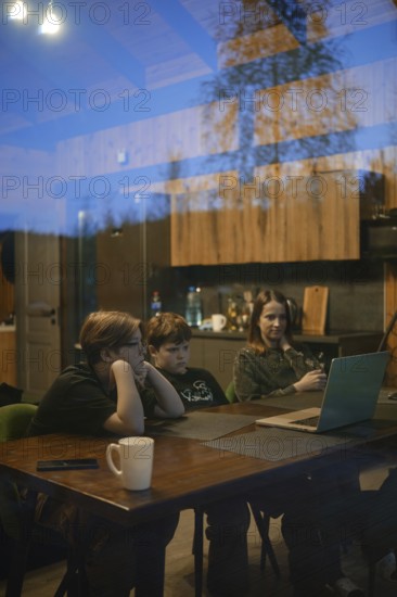 A cozy setting captures two children and an adult focused on a laptop screen inside a modern home. The warm wood interior reflects a comfortable atmosphere during the evening