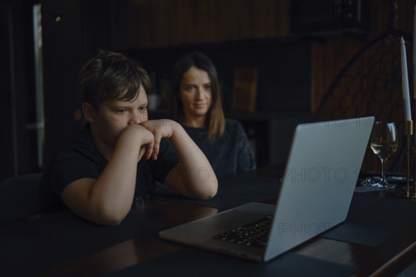 A boy sits focused at a table, watching a laptop screen closely while his mother looks on with a supportive expression