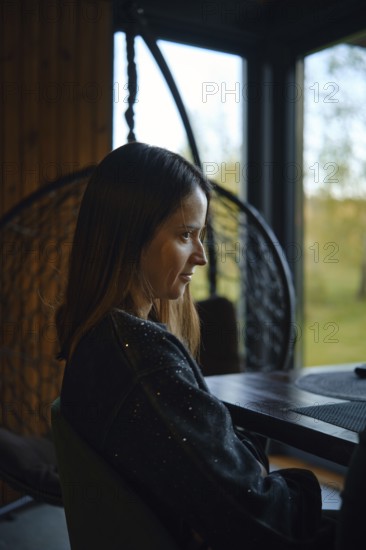 A woman is seated at a table in a warm, inviting indoor space with large windows. The setting is peaceful, filled with natural light from the golden hour, creating a serene atmosphere