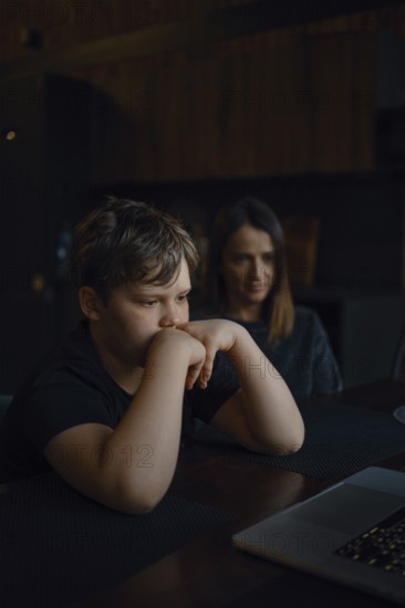 A young boy sits at a table, staring intently at a laptop screen with his hands on his chin. His mother watches him closely from behind, both in a cozy, dimly lit room