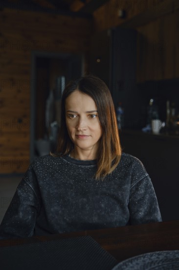 A woman with medium-length brown hair sits at a dark wooden table, reflecting thoughtfully. The rustic cabin atmosphere is warm with natural wooden elements and a soft light filtering in