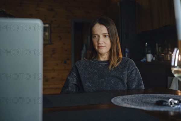 A woman is focused on her laptop while sitting at a wooden table in a warm dark living room. The cozy interior features a rustic design, enhancing her home office atmosphere