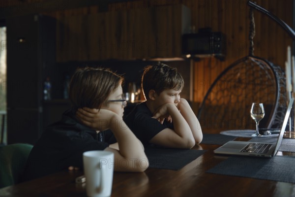 Two children are seated at a wooden table in a warm, rustic environment, deeply engaged with a laptop screen