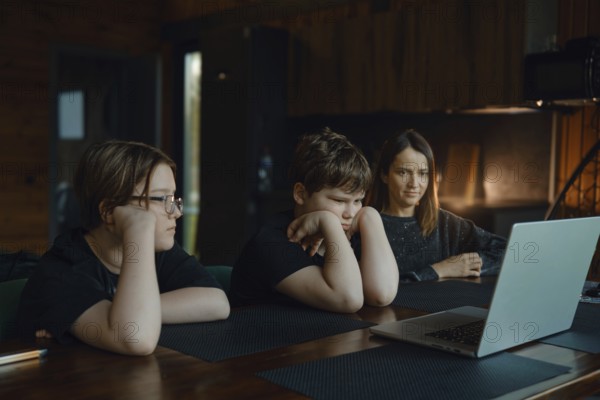Two children and their mother sit at a wooden table, watching a laptop screen. The room is warm and inviting, with soft lighting and natural wood accents