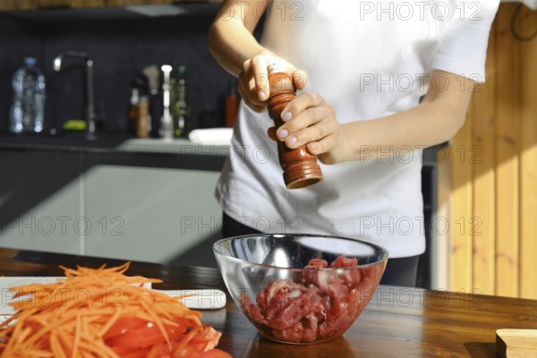 Unrecognizable person seasons fresh beef meat with pepper while preparing a healthy meal in a bright kitchen. Colorful vegetables like carrots sit nearby, ready to be included in the dish