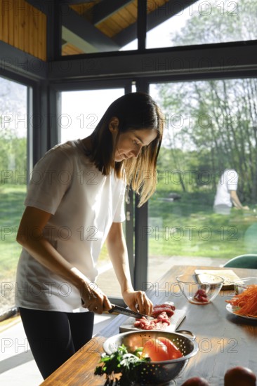 A woman is slicing beef meat in a sunlit kitchen with large windows. Fresh produce is spread on the table, showcasing a meal preparation session filled with vibrant colors and healthy choices