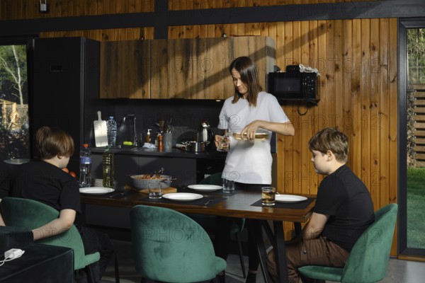 In the kitchen, a woman pours a drink, while two children are sitting at the dining table, impatiently waiting for their meal. The cozy atmosphere demonstrates a family pastime focused on healthy eating