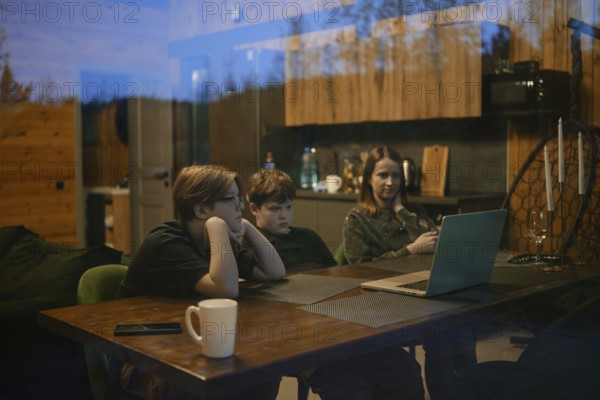 Inside a cozy cabin, a family gathers around a table enjoying a shared experience. Two children are focused on a laptop, while an adult casually checks a phone, creating a warm atmosphere