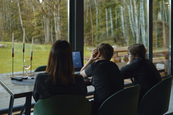 In a cozy lodge surrounded by nature, a family gathers at a table. Two children sit in front of a laptop, focused on their screen. An adult watches them, creating a warm family moment
