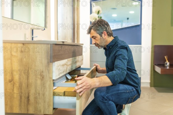 Mature man crouching, opening drawers of a bathroom vanity, checking quality and organization in a home improvement store showroom