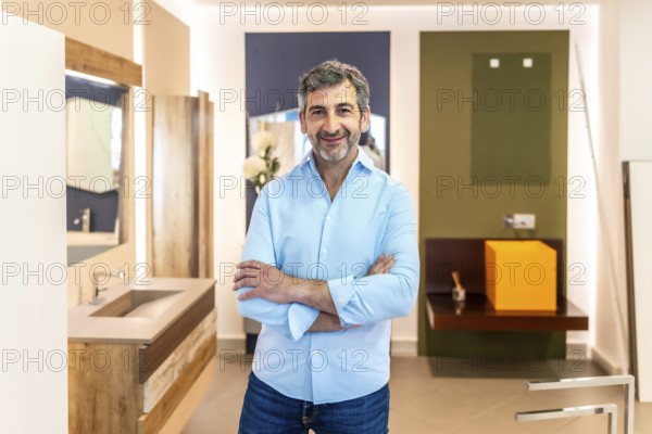 Confident salesman smiling with crossed arms while posing in a bathroom furniture store, showcasing his expertise and welcoming customers