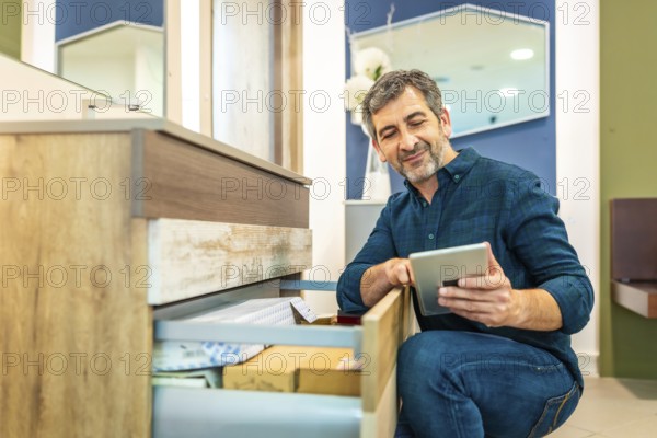 Smiling mature man using a digital tablet while checking out furniture options in a bathroom furniture store, enjoying the shopping experience