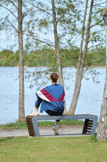A woman sits on a unique bench facing a serene lake, surrounded by trees. She enjoys the tranquil setting, soaking up the calm atmosphere during a sunny day. Nature's beauty enhances the moment