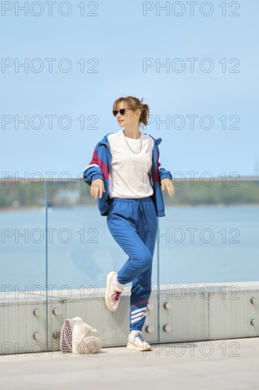 A woman relaxes by a glass railing at the waterfront on a bright sunny day. She wears a colorful sporty outfit and sunglasses, exuding a casual and trendy vibe while enjoying the view