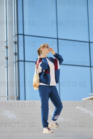 A young individual stands on a staircase, wearing sporty clothing and sunglasses. The sunny weather complements the vibrant blue building behind, creating a lively urban atmosphere