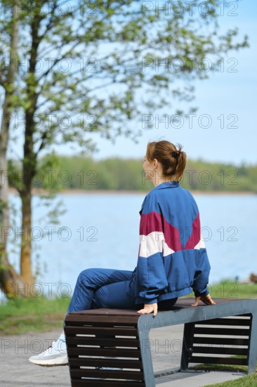 A person sits on a bench by a serene lake surrounded by lush greenery, enjoying the calm atmosphere. It is a bright spring afternoon, perfect for unwinding outdoors