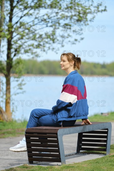 A young woman relaxes on a modern bench near a serene lake, dressed in colorful activewear. The bright sun and blue sky enhance the tranquil atmosphere, highlighting her joy in nature