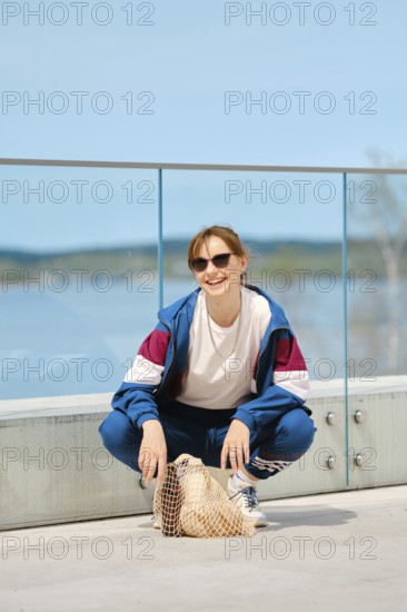 A young person squats on a modern deck by a scenic body of water, wearing sunglasses and a sporty outfit