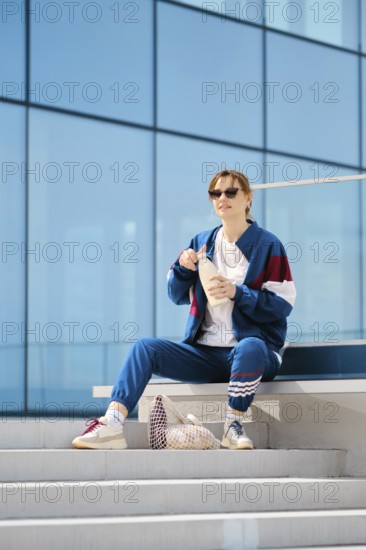 A young woman dresses in a stylish tracksuit sits on outdoor steps near a contemporary structure. They savor a snack from a container and wear sunglasses, basking in the warm sunlight