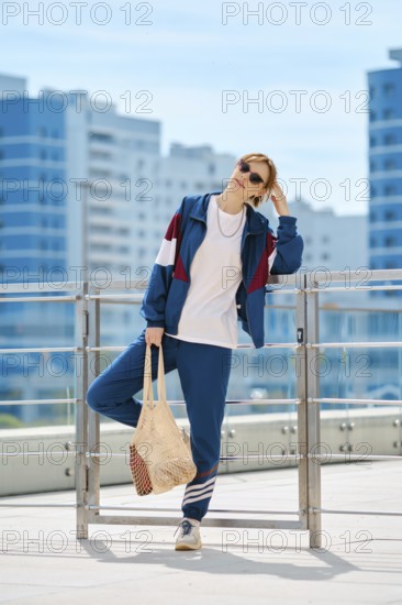 A woman dressed in a sporty outfit stands confidently on a terrace surrounded by a bright blue sky and modern buildings