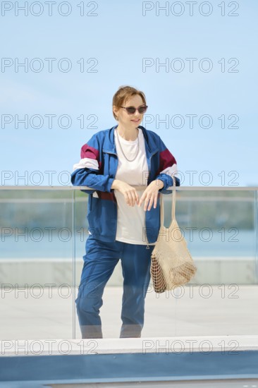 A woman stands by a glass railing at a waterfront location, dressed in a colorful jacket and sunglasses. She sports a relaxed look, holding a netted bag as she takes in the scenery