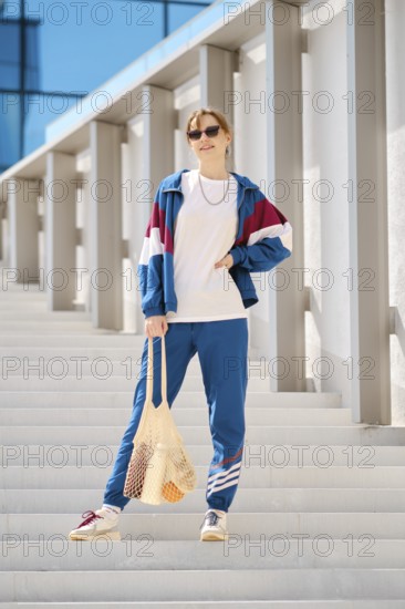 A young woman stands on a series of outdoor steps, wearing a colorful sports outfit with sunglasses. She holds a reusable string bag with food