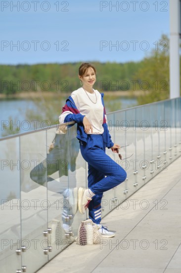 A woman dressed in comfortable sportswear leans casually against a glass railing overlooking a serene body of water