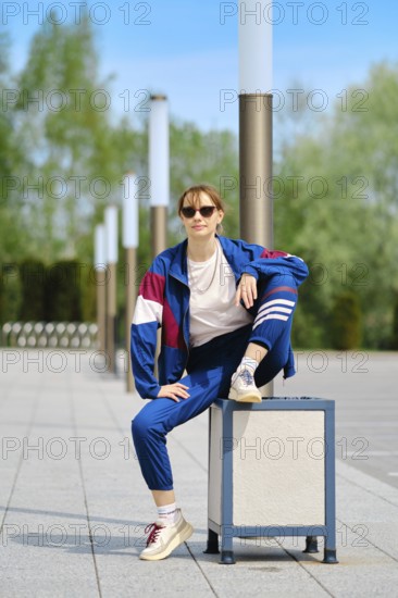 A woman dressed in a blue and maroon sporty outfit sits on a trashcan with one leg raised, showcasing a relaxed pose. The urban park features green trees and clear blue skies in the background