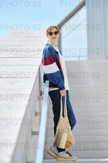 A young sporty woman with string bag stands on a staircase, dressed in stylish athletic track suit