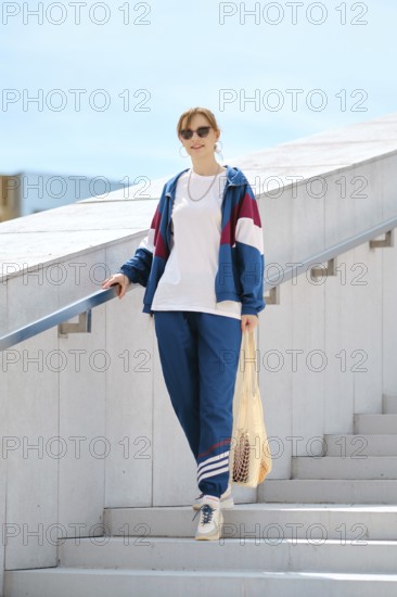A young woman with sunglasses descends a staircase wearing a colorful sporty outfit. She carries a the string bag. The bright daylight enhances the casual, relaxed atmosphere of the setting