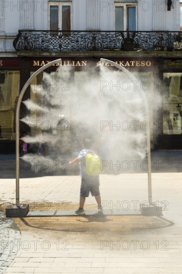 A mobile spray arch sprays a fine mist of water in a sunny square. A child uses the refreshment in the summer heat, Bratislava, Slovakia