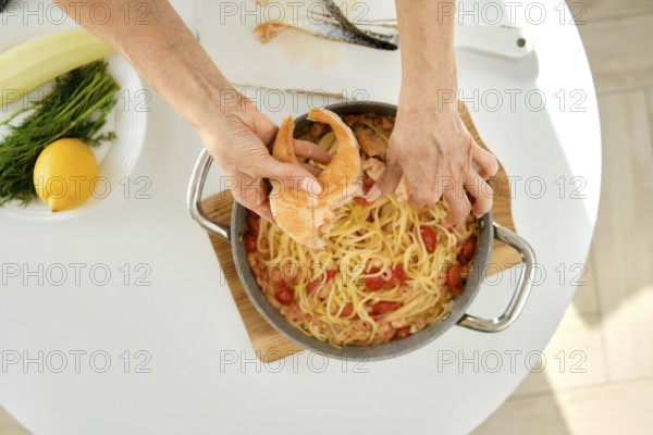 A person is adding salmon to a pot of pasta with tomatoes and herbs on a sunny kitchen countertop. Fresh ingredients like lemon and greens are nearby, indicating a home-cooked meal