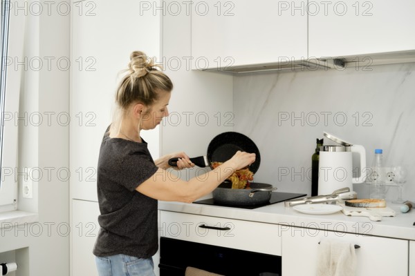 A woman stands in a bright, modern kitchen, carefully stirring a pan filled with colorful ingredients as she prepares a meal
