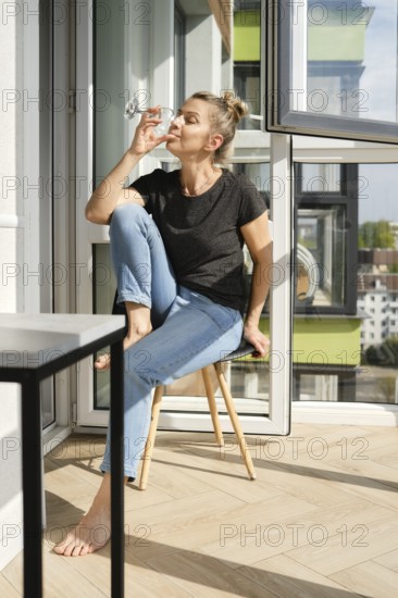 A middle-aged woman sits comfortably on a wooden chair in a sunny balcony, sipping from a glass. Natural light streams in, highlighting her casual style and the greenery of the city
